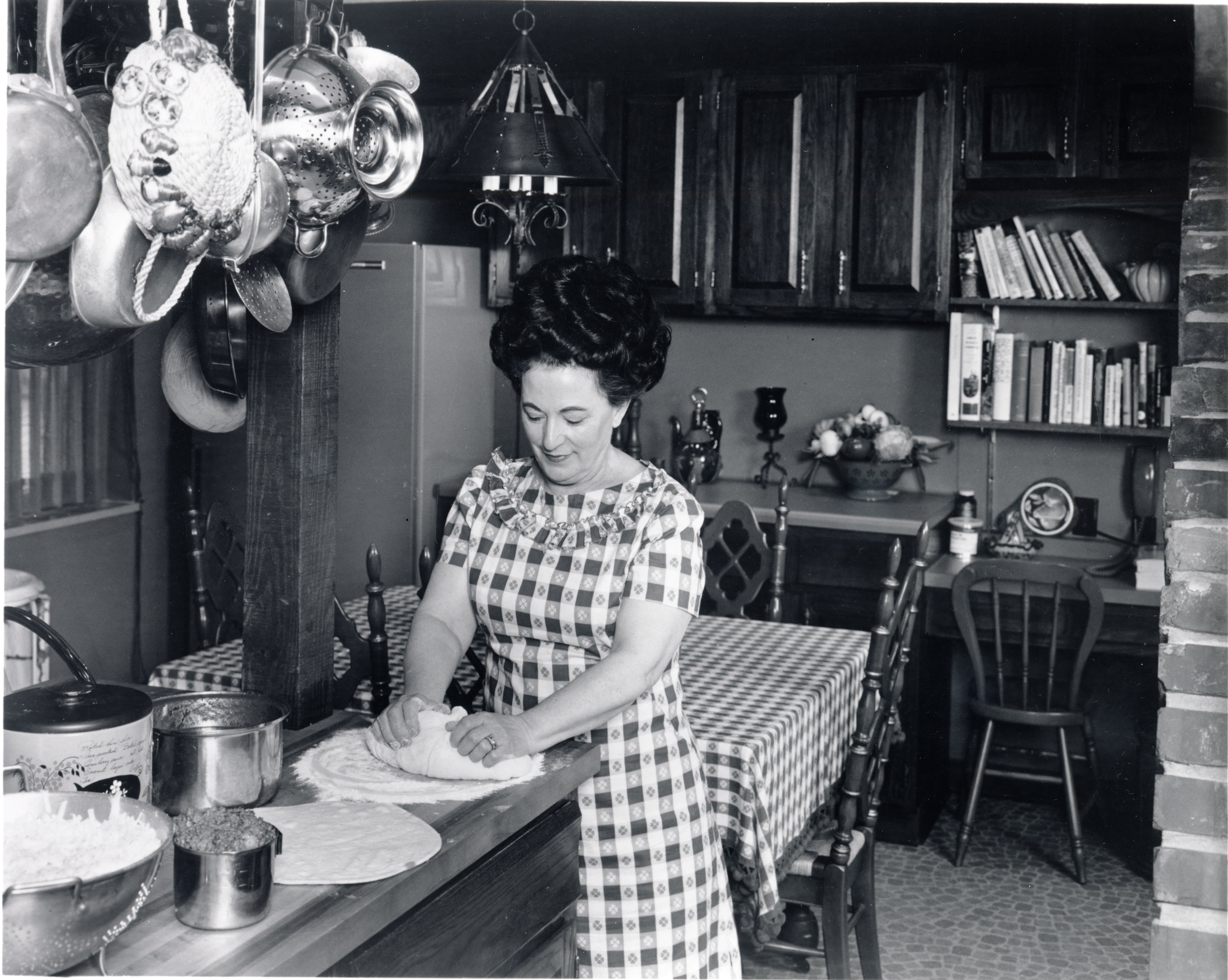 Black and white photo of Rose Totino wearing a checkered dress while hand kneading homemade pizza dough on her kitchen counter.