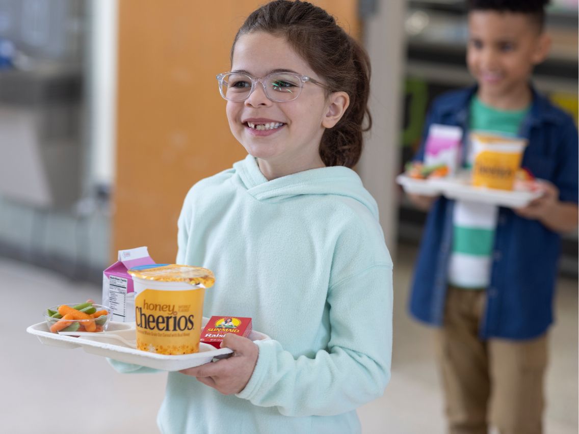 A smiling child holds a cafeteria tray containing Honey Cheerios, a milk carton, and small container of carrots and sugar snap peas. Another child in the background holds a similar tray. The setting appears to be a school cafeteria.